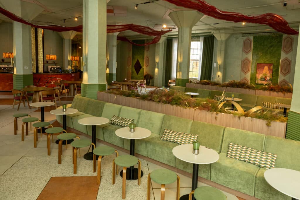 Zona lounge con sofás verdes, mesas redondas y vegetación entre columnas