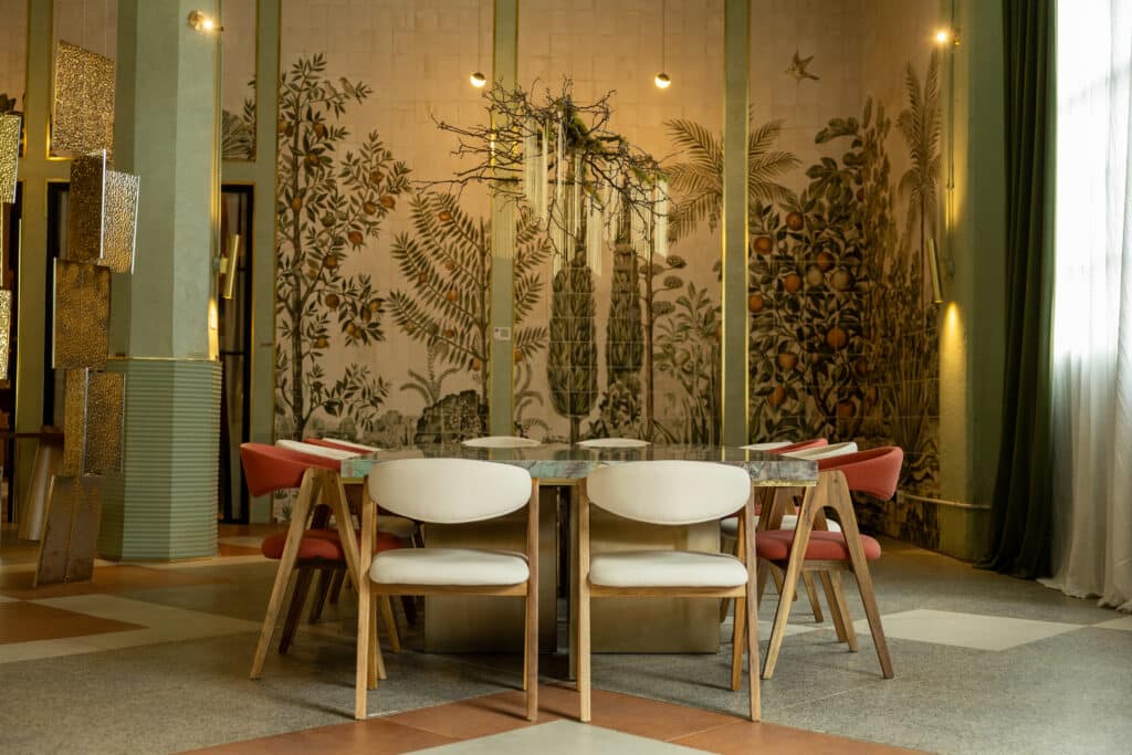 Mesa de comedor con sillas claras y rojas frente a un mural vegetal y una luminaria de ramas